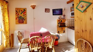 A cozy kitchen with wooden cabinets, a dining table with chairs, a TV mounted on the wall, and a window with curtains.