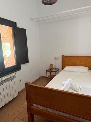 A bedroom with a wooden bed, a small bedside table, a radiator, and a window with a view of greenery. - MASOVERS A, 3 bedrooms, 1–6 people (max. 8—2 on a sofa bed), 5 min. from the beach (Castelló d'Empúries)