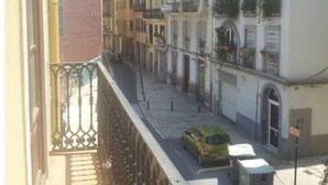 Eine schmale Straße mit Häusern an beiden Seiten, darunter Balkone mit Pflanzen und ein grünes Auto, das am Straßenrand geparkt ist.
