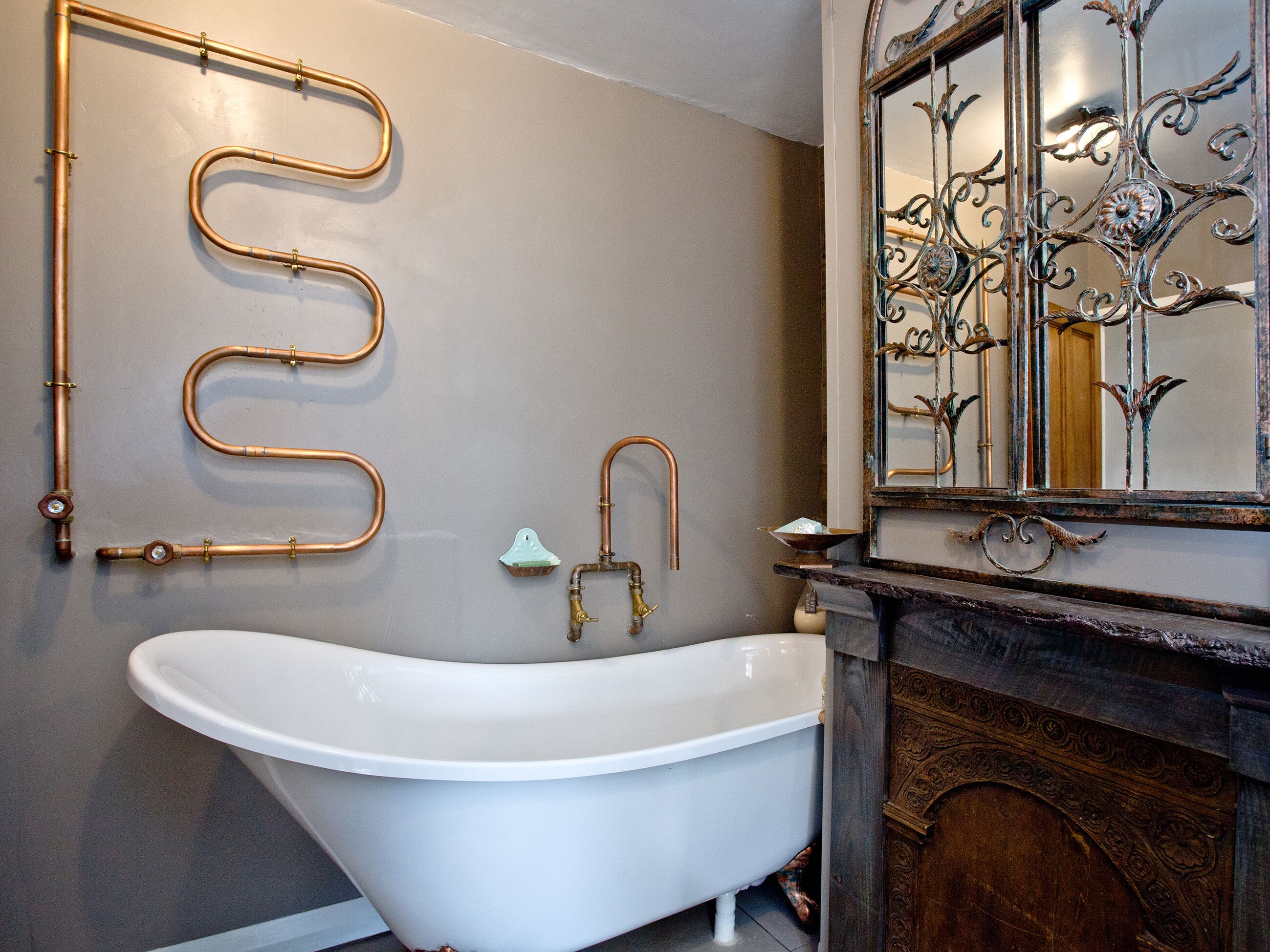 Un baño con una bañera independiente, tuberías de cobre en la pared, un jabonera en forma de pájaro pequeño y un espejo decorativo con ornamentación metálica.