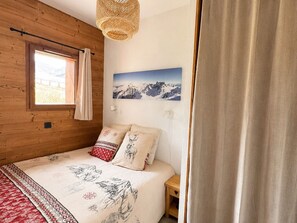 A bedroom with a wooden wall, a bed with patterned bedding, a window with curtains, a wall-mounted picture of mountains, and a bedside table.