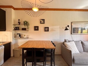 A compact kitchen with a wooden table and chairs, a built-in oven, and a sink. There is a grey sofa with cushions and a framed picture on the wall.