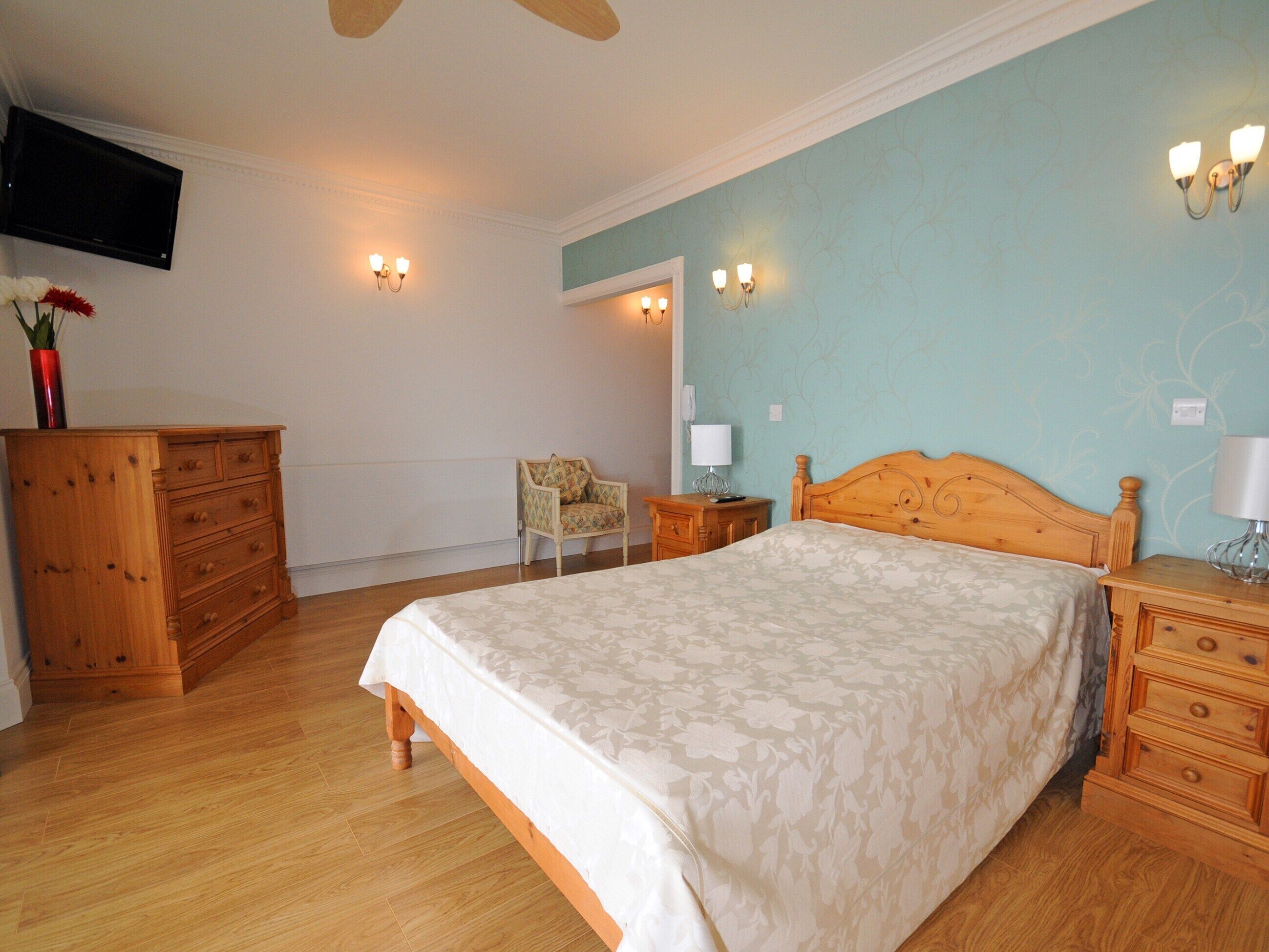 A bedroom with a bed, wooden dresser, chair, nightstands, and a television on the wall.