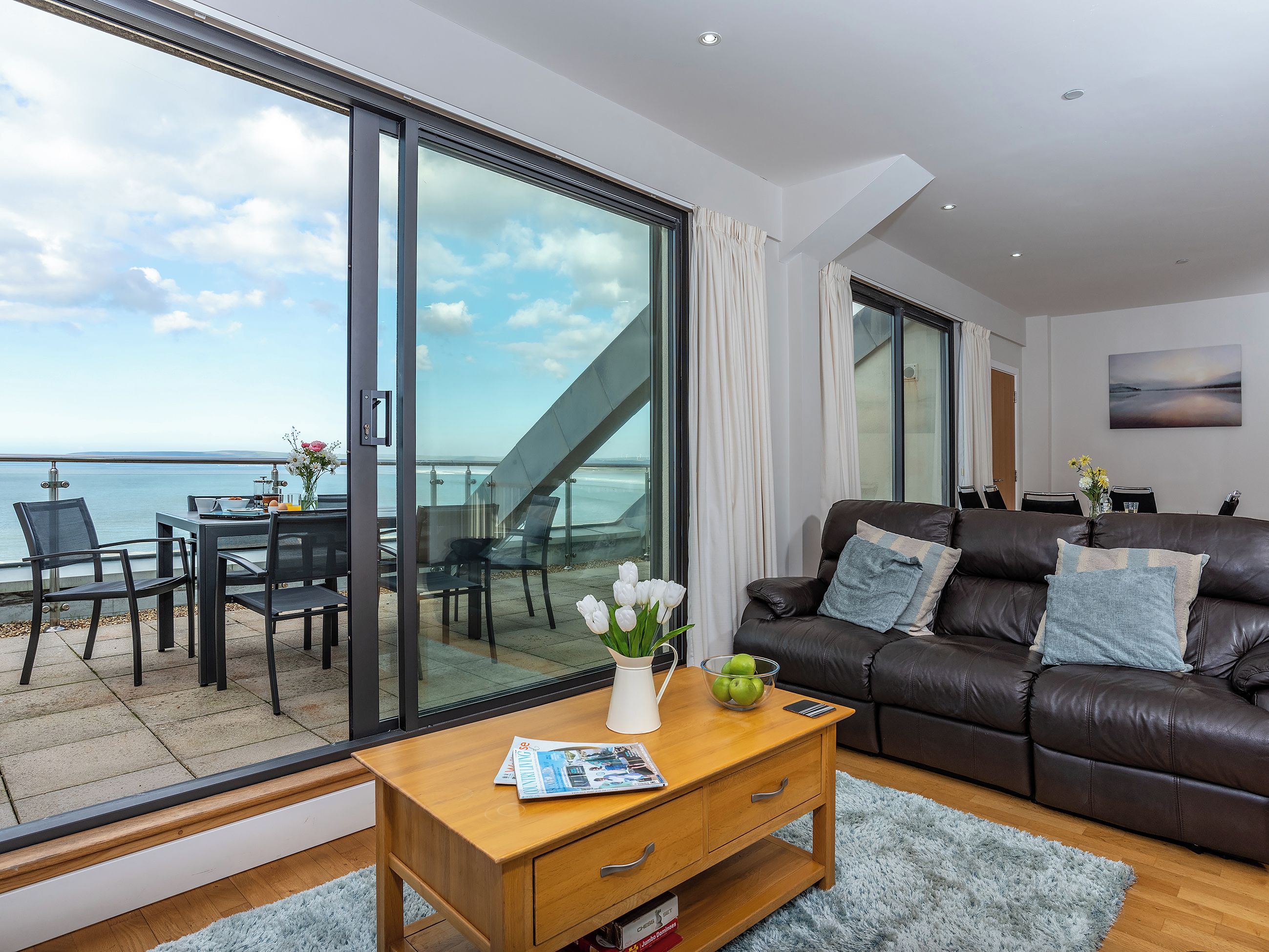 A modern living room with a large sliding glass door leading to a balcony overlooking the sea.
