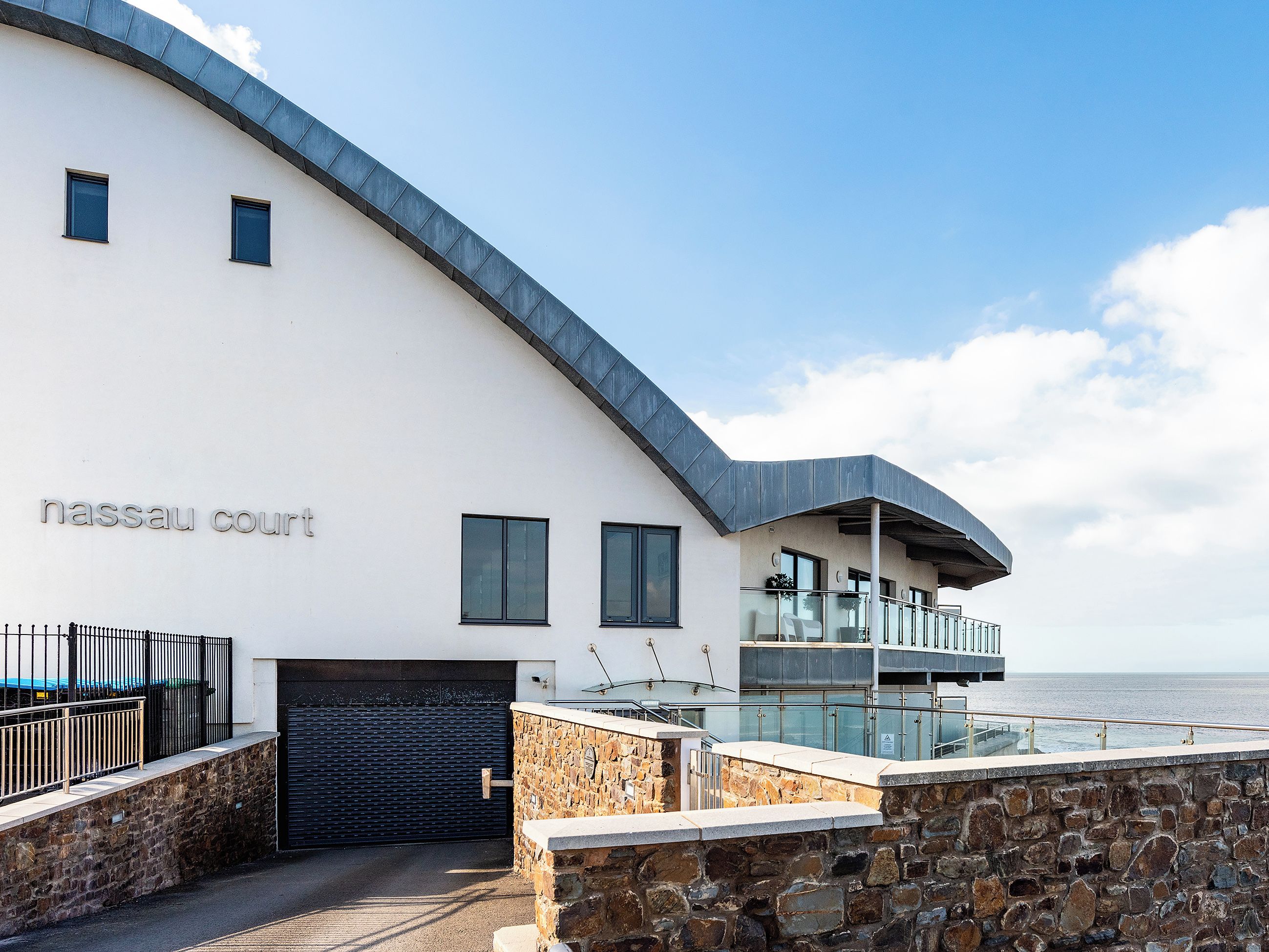 A modern building with a curved roof, a stone wall, and a glass balcony overlooking the sea.