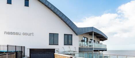 A modern building with a curved roof, a stone wall, and a glass balcony overlooking the sea.