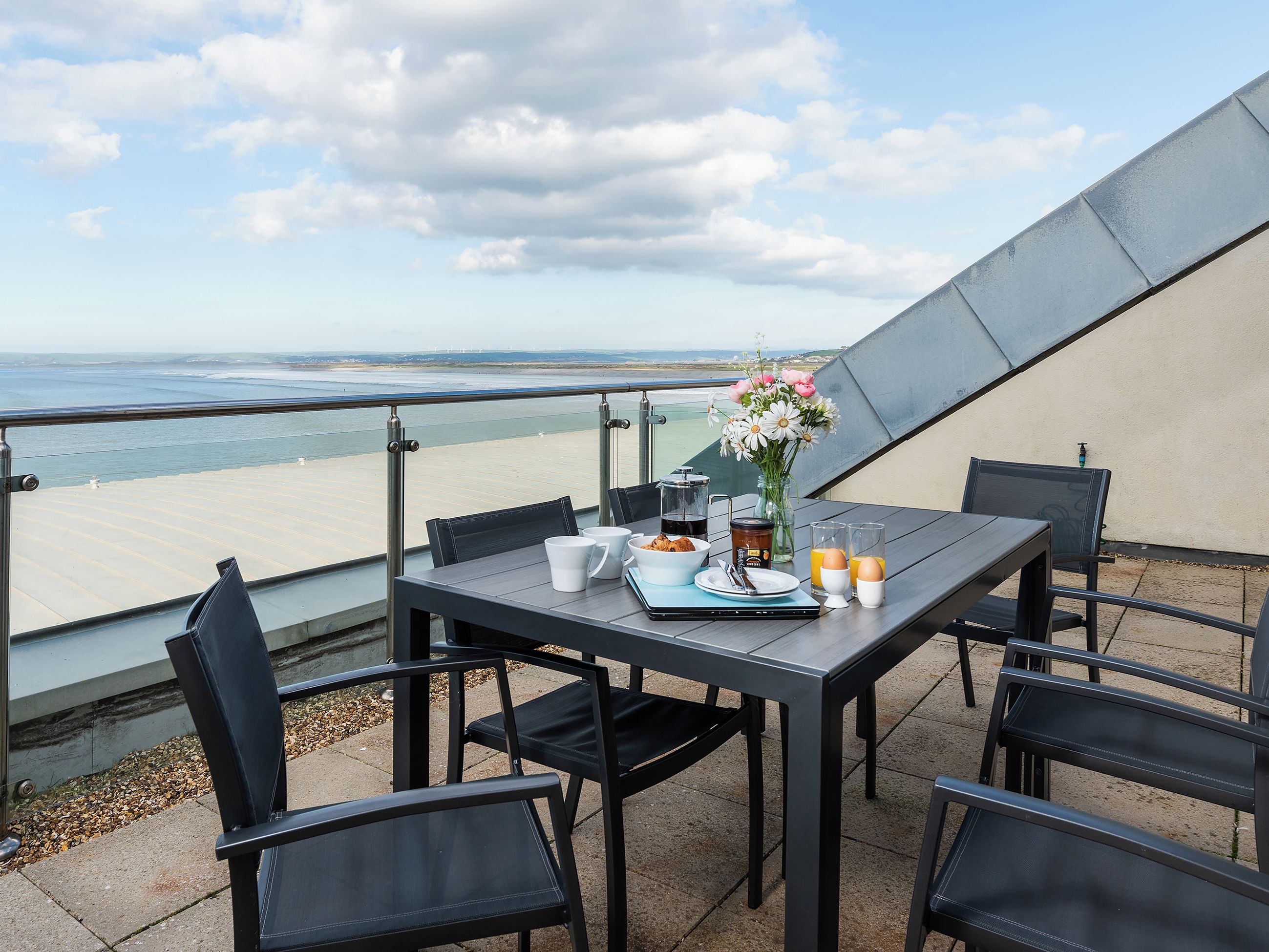 A table with breakfast items, a vase of flowers, and a view of the beach.