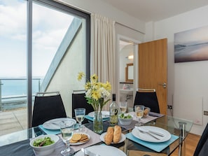 Una mesa de comedor dispuesta con platos, vasos y un florero, con vistas a un balcón y al mar.