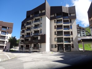 A multi-story residential building with balconies, a parking area, and a clear sky.