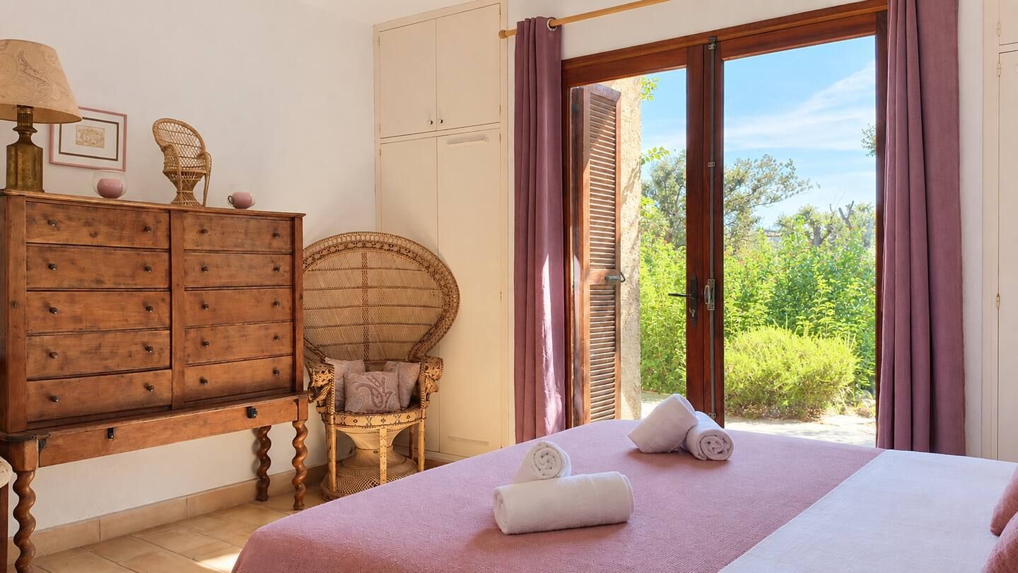 Un dormitorio con una cama, un aparador de madera, una silla de mimbre y vistas a la vegetación a través de contraventanas abiertas.