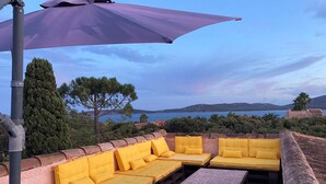 A terrace with yellow cushioned seating, a purple umbrella, and a view of the sea and hills. - LARGE VILLA, SOUTH CORSICA, PRIVATE AREA OF CALA ROSSA. (Lecci)