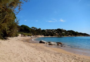 A sandy beach with clear blue water, rocky outcrops, and a forested hillside in the background.