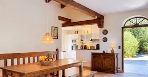 A wooden dining table with a fruit basket, a bench, a pendant light, and a view into a kitchen area with cabinets and a window. - LARGE VILLA, SOUTH CORSICA, PRIVATE AREA OF CALA ROSSA. (Lecci)