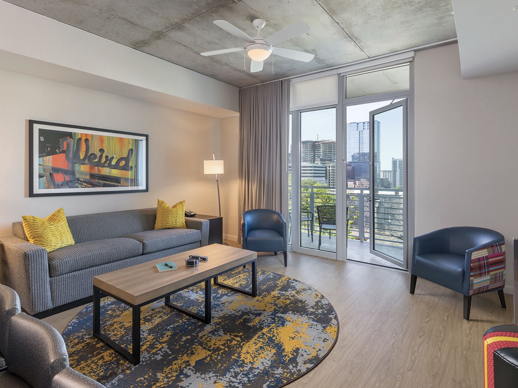 A modern living room with a grey sofa, a coffee table, and a blue armchair. There is a large window with a view of the city.