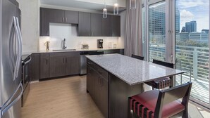 A modern kitchen with dark cabinets, a granite countertop, and a large island. There is a dining area with a red chair and a view of the city through large windows. - Downtown Austin 2 Bedroom Deluxe with Rooftop Pool in the Heart of everything (Austin)