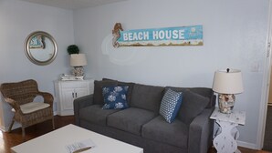 Una sala de estar con un sofá gris, una silla de mimbre, una mesa blanca y un letrero en la pared que dice "BEACH HOUSE".