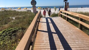 Pasarela de madera que lleva a una playa con sombrillas y gente caminando.