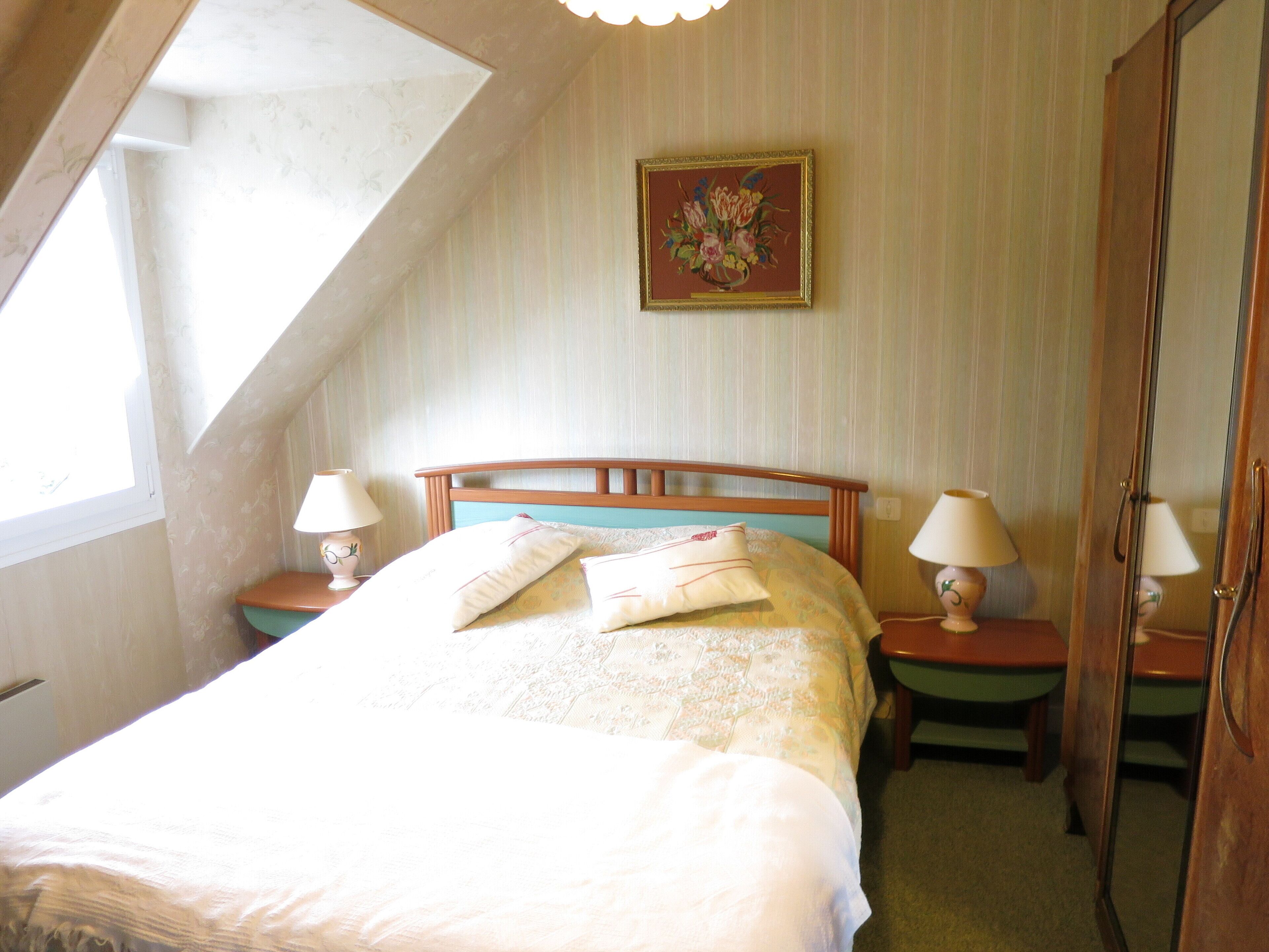 Een slaapkamer met een schuine kap, een bed met kussens, twee bedlampen, een spiegel en een bloemen schilderij aan de muur.