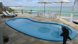 A swimming pool with a dolphin sculpture, lounge chairs, and umbrellas overlooking a beach with boats. - Pousada Ecobaía (Baia Formosa)