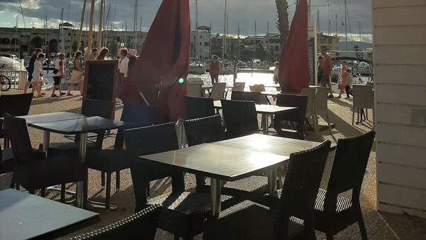 Et udendørs opholdsområde med borde og stole, med udsigt over en lysthavn med både og en klar himmel.