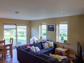 A living room with a sofa, a small table, a lamp, and a television. There are windows with a view of the outdoors.
