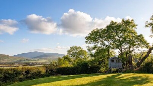 Paisagem rural com uma pequena casa branca cercada por vegetação e uma cordilheira ao longe.