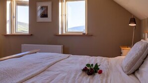 Quarto com teto inclinado, duas janelas, cama com edredom branco e um buquê de flores vermelhas, abajur e um quadro na parede.