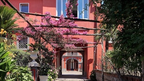 A courtyard with a red building, a wooden pergola with pink flowers, and a white column. - Sophia - Lovely Apartment   (Meta)