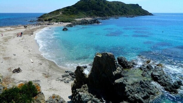 A beach with clear turquoise water, rocky outcrops, and a small hill in the background.