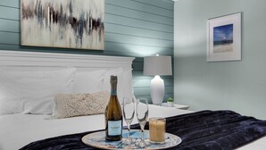 Uma cama bem arrumada com cabeceira branca, um ornamento de parede decorativo, um quadro emoldurado e uma mesinha de cabeceira com abajur e uma pequena planta.