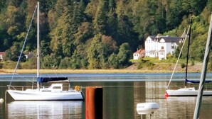 Un lac paisible où flottent des bateaux, une maison perchée sur une colline et, au loin, une forêt dense.
