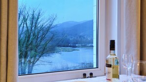 Une table à manger avec un saladier, une bouteille de vin et des verres, une fenêtre donnant sur une rivière et des montagnes.