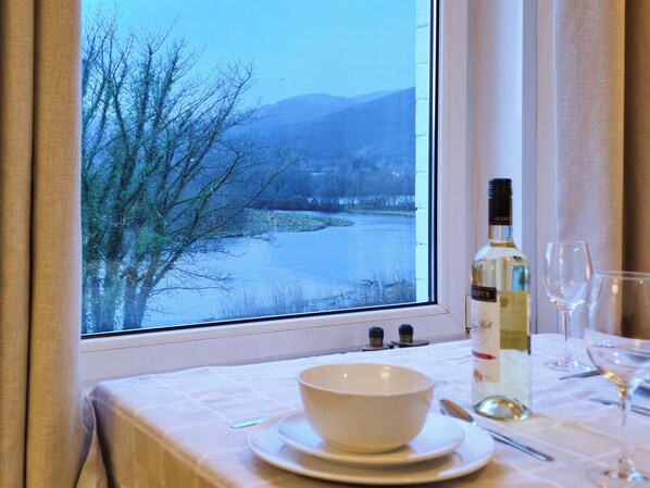 A dining table with a bowl, wine bottle, and glasses, a window with a view of a river and mountains.