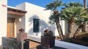 A modern building with a white exterior, a wooden door, and a stone pathway leading to a door. There are two potted plants with red flowers on the pathway. - Luxury villas Mamma Mía Villa A Parking & pool private (Playa Blanca)