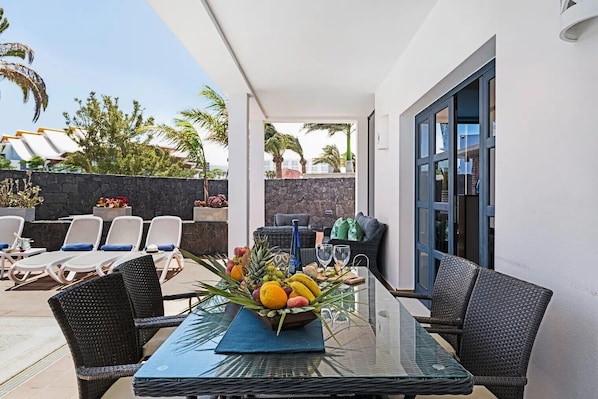 A sunlit outdoor patio with a glass table, wicker chairs, and a fruit centerpiece.