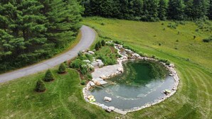 An aerial view of a landscaped area with a circular pond, a winding road, and a variety of greenery.
