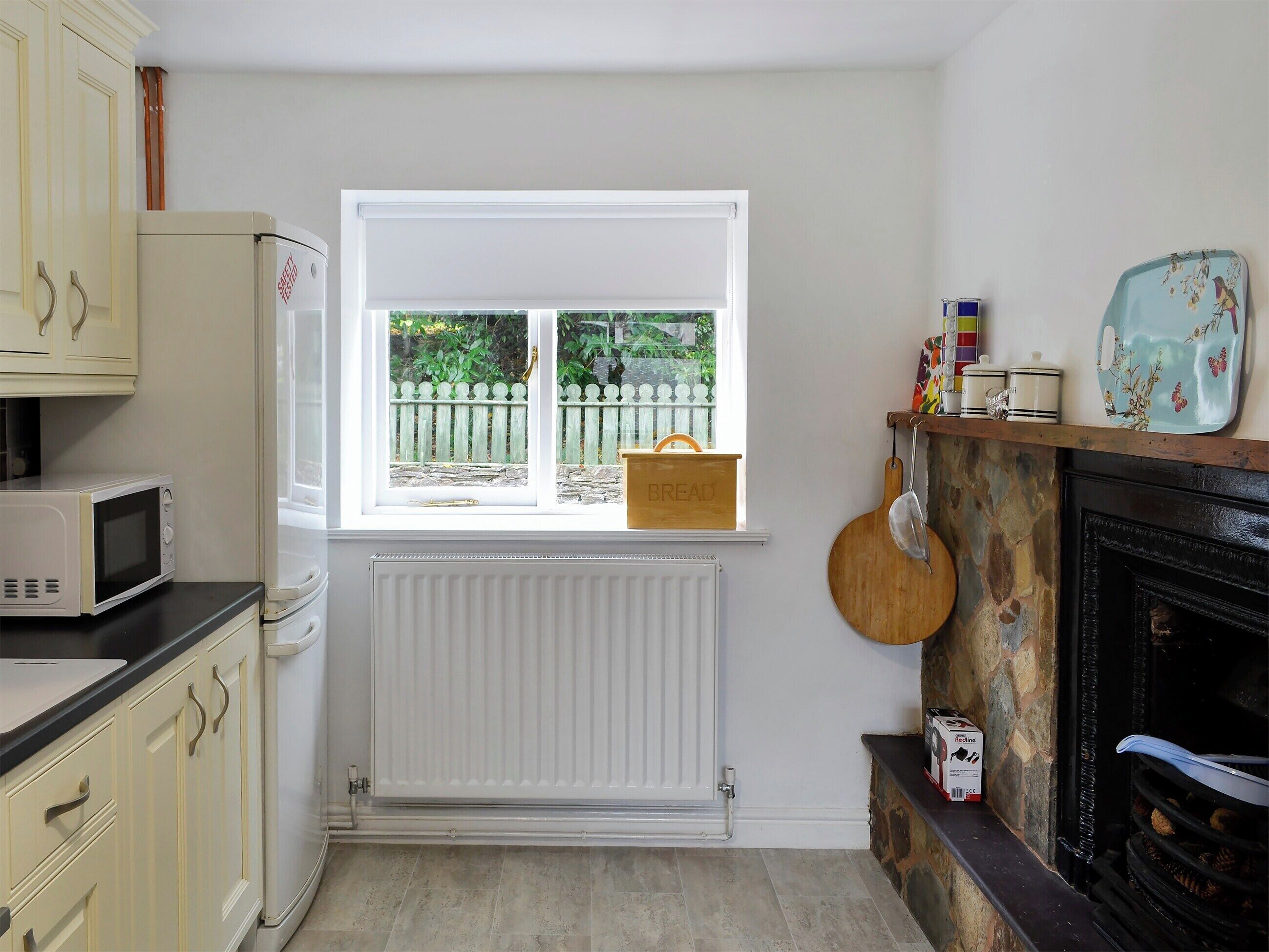 Een keuken met een witte radiator, een magnetron, een raam met uitzicht op een groene schutting, en een open haard met een stenen schoorsteenmantel.