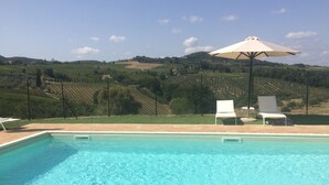 A swimming pool with clear blue water, surrounded by a patio area with two white lounge chairs and a large umbrella. In the background, there are rolling hills and vineyards.