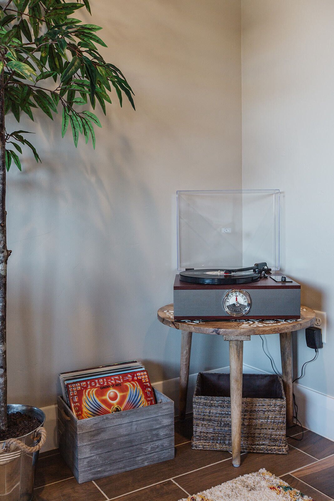 A corner room with a potted plant, a record player on a wooden table, and a vinyl record box.