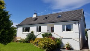 A two-story house with a white exterior, a grey roof, and a chimney. There are two windows on the first floor and one on the second floor. The house is surrounded by a well-maintained garden with various plants and a lawn. - Dunshee (Dunvegan)
