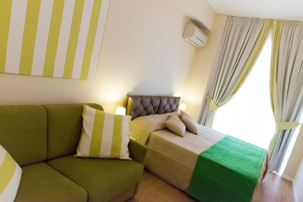 Een kamer met een groene bank, een bed met een grijze hoofdbord en een raam met gordijnen.