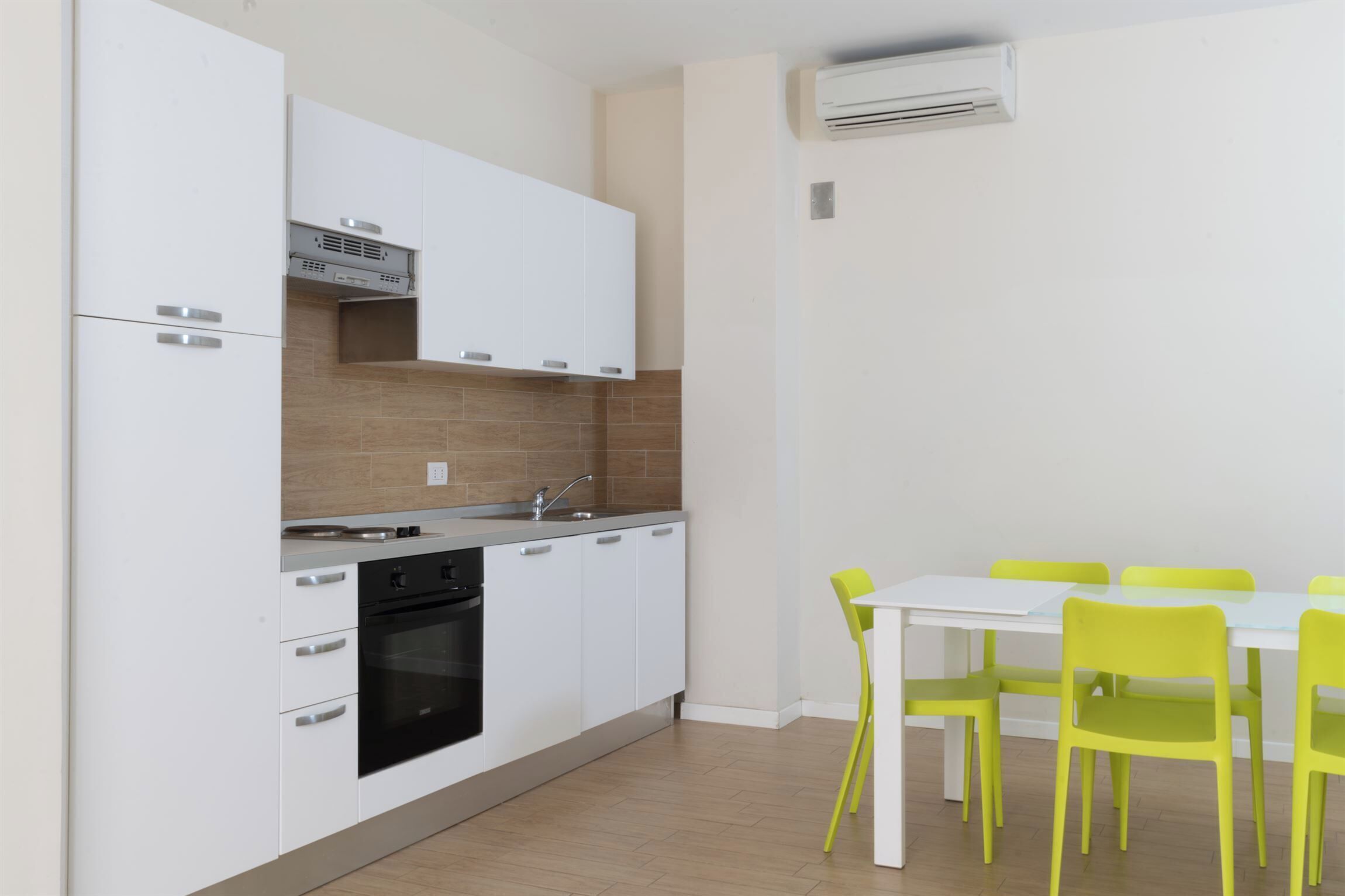 Une cuisine moderne avec des armoires blanches, un four encastré et un coin repas comprenant une table blanche et des chaises jaunes.