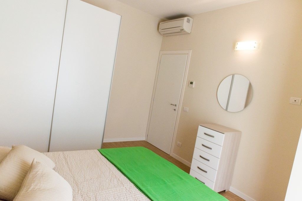 A bedroom with a bed, a wardrobe, a dresser, an air conditioning unit, and a wall-mounted mirror.
