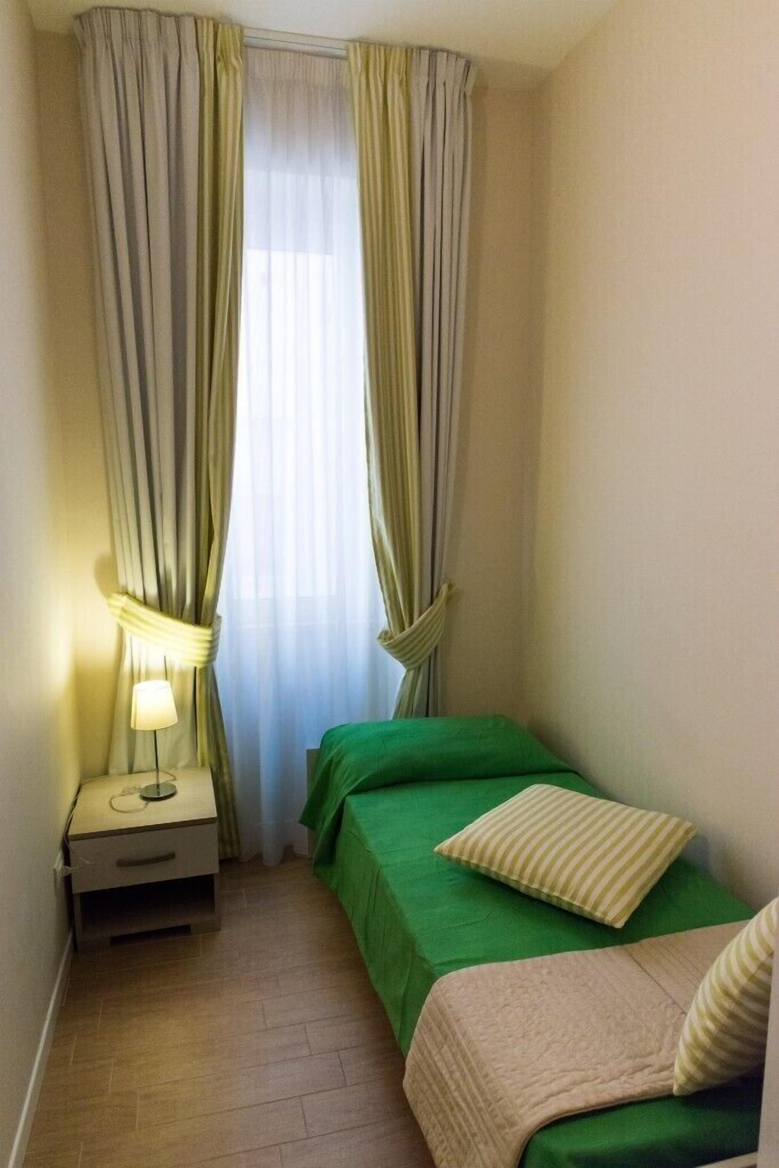A small, neatly arranged room with two beds, a bedside table with a lamp, and a window with curtains.