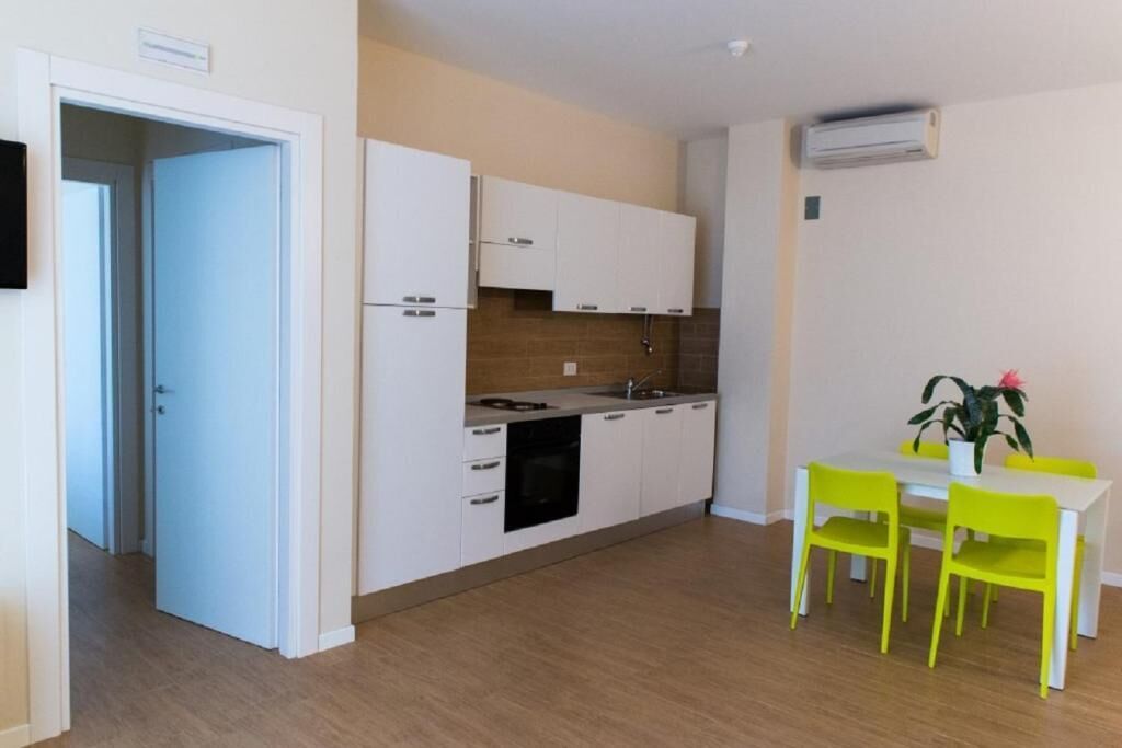 Uma cozinha compacta com armários brancos, forno embutido e pia. Há uma mesa de jantar com duas cadeiras e um vaso de planta.