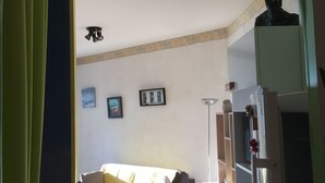 Een woonkamer met een bank, een bijzettafeltje en een koelkast. Er is een doorgang naar een andere kamer.