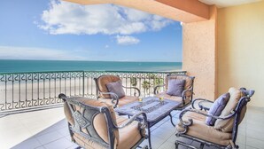 A balcony with two cushioned chairs and a table, overlooking a beach and ocean.