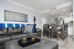 A modern living room with a grey leather sofa, a coffee table with two glasses of whiskey, and a dining area with wooden chairs and a kitchen in the background.