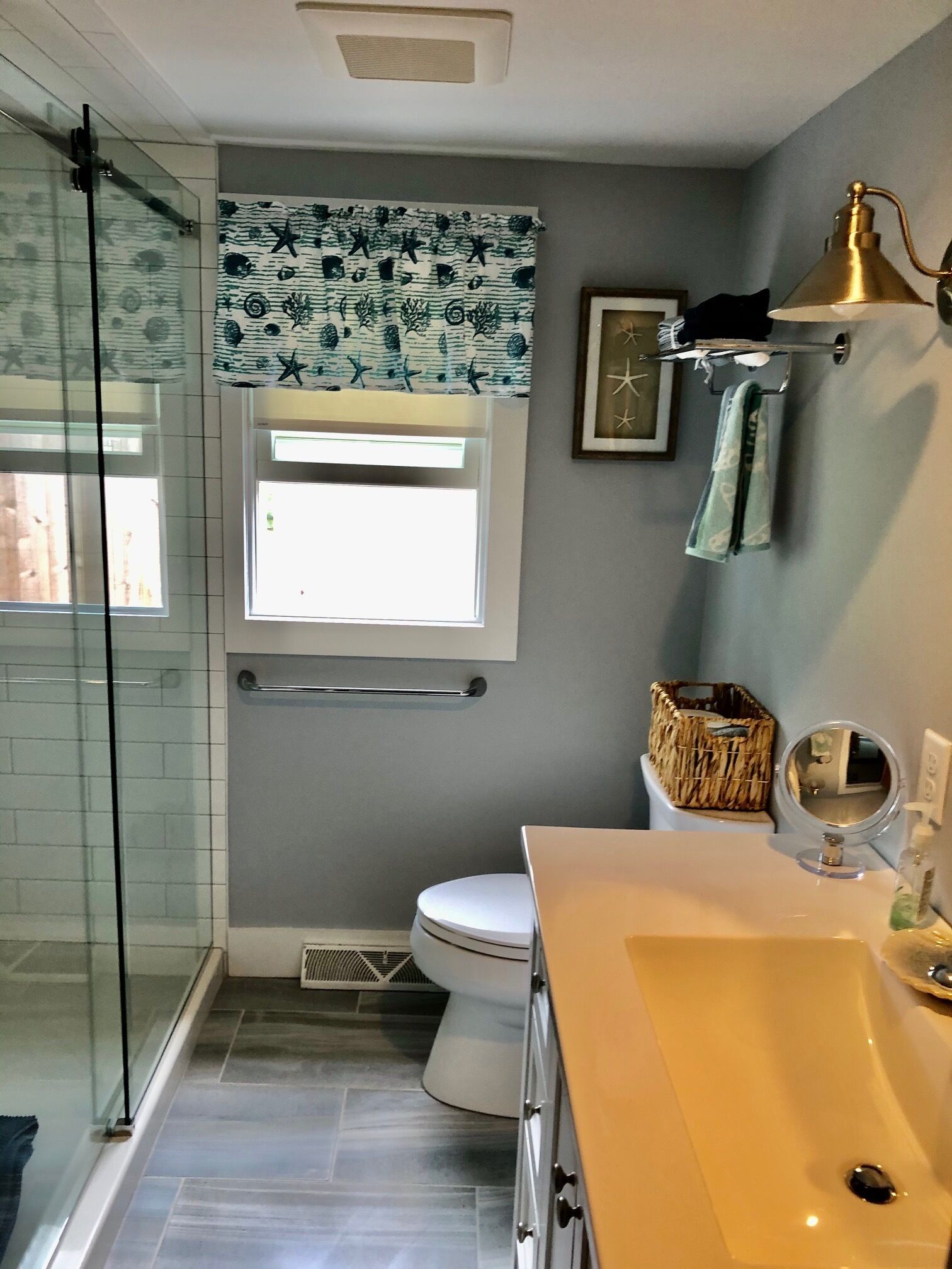 A bathroom with a glass shower enclosure, a white toilet, a vanity with a mirror, and a window with a patterned curtain.