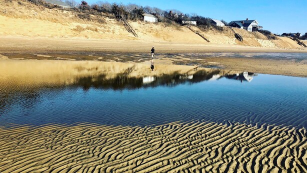 Ein sandiger Strand mit ruhigem Gewässer, das Gebäude spiegelt, und eine Person.